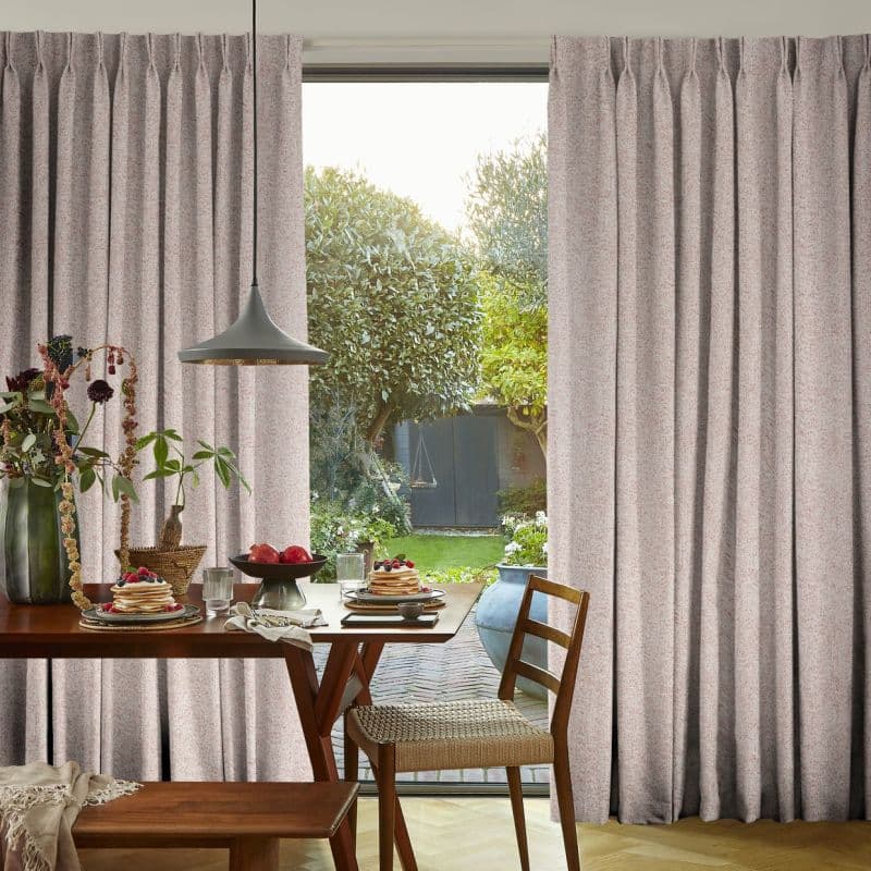 Full-length pale pink textured pinch-pleat curtains, drawn aside to reveal a glass door; soft daylight falls on a wooden dining table set for breakfast with pancakes and wooden chairs.