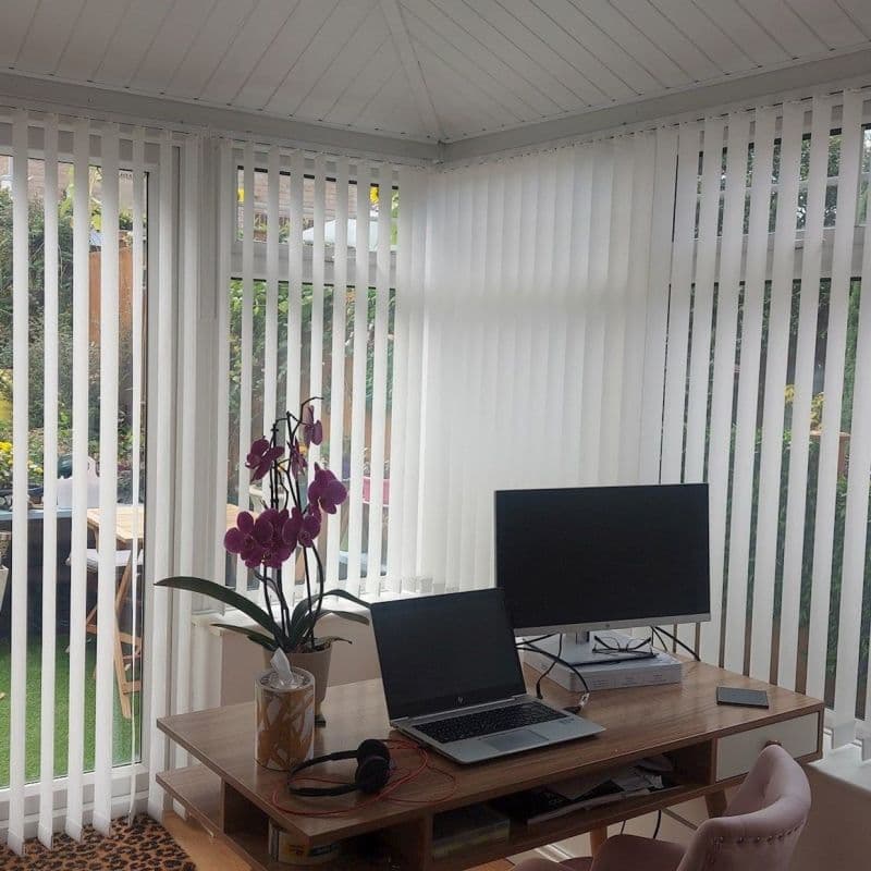 White vertical blinds across wraparound conservatory windows, straight slatted and mostly closed, filtering soft daylight into a home office; wooden desk with laptop, monitor, orchid and pink chair, garden outside.