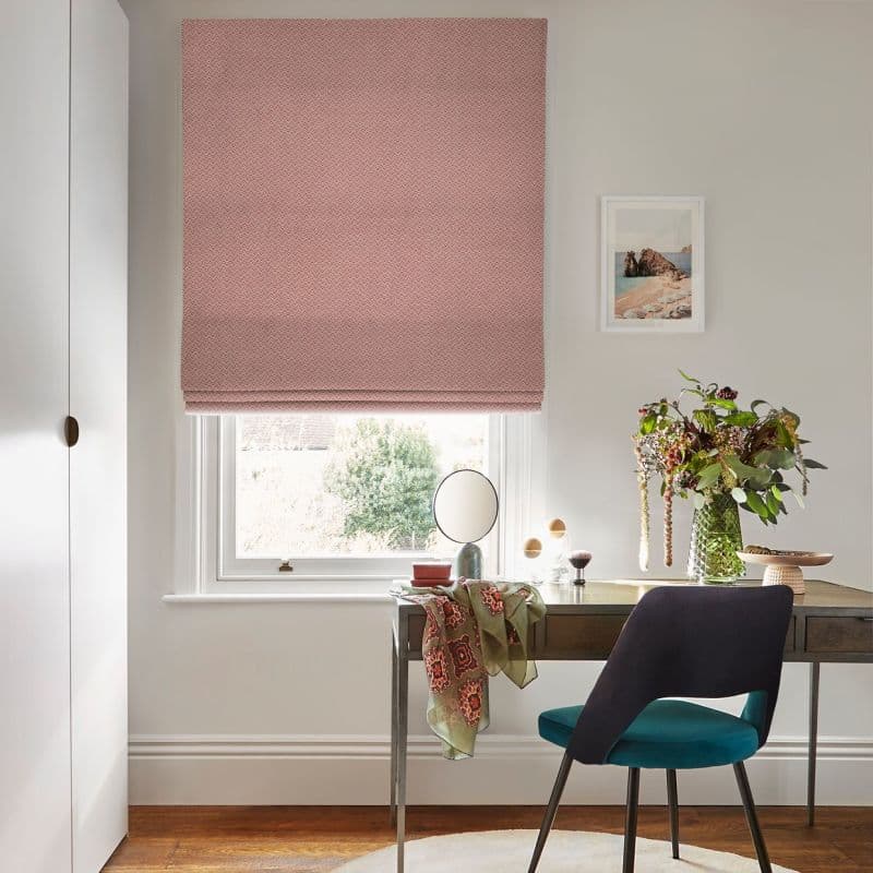 A pink patterned Roman blind lowered nearly fully over a sash window, filtering soft daylight; a desk with mirror, chair and vase of flowers beneath in a bright home office.