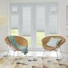 Pleated light-grey blinds on French doors and sidelights, partially lowered and filtering daylight, revealing lawn below; bright living room with two wicker chairs, pastel cushions, geometric rug and floor lamp.
