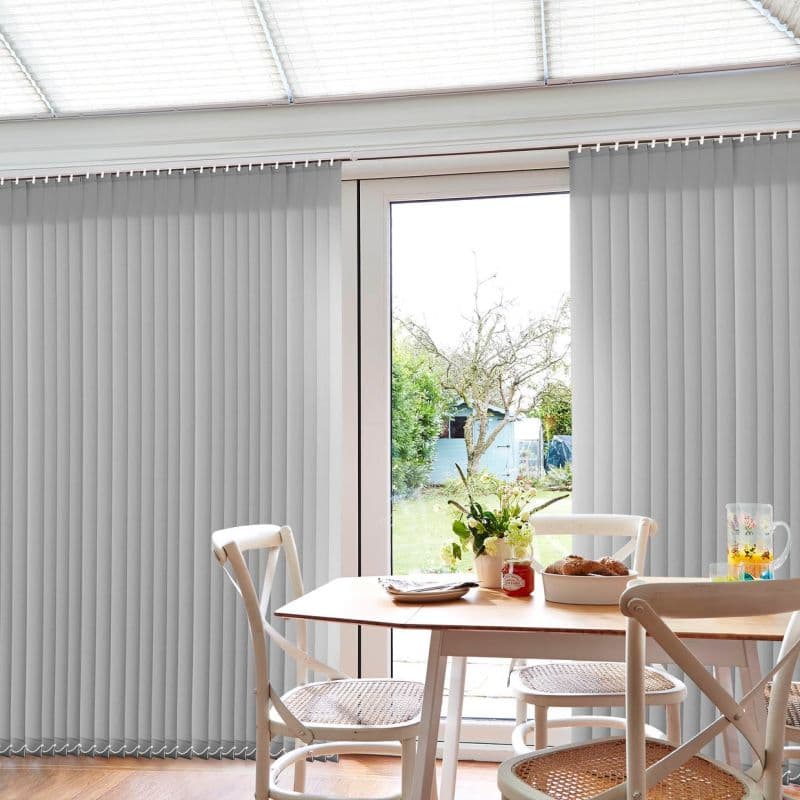 Light-gray vertical blinds cover a sliding glass door; slats hang vertically and are parted centrally, filtering bright daylight into a small dining area with table, chairs, vase, and garden view.