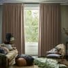 Full-length pleated beige curtains, hung on a ceiling track, mostly closed with a narrow central opening, framing and filtering daylight from a leafy garden visible through a living-room window.