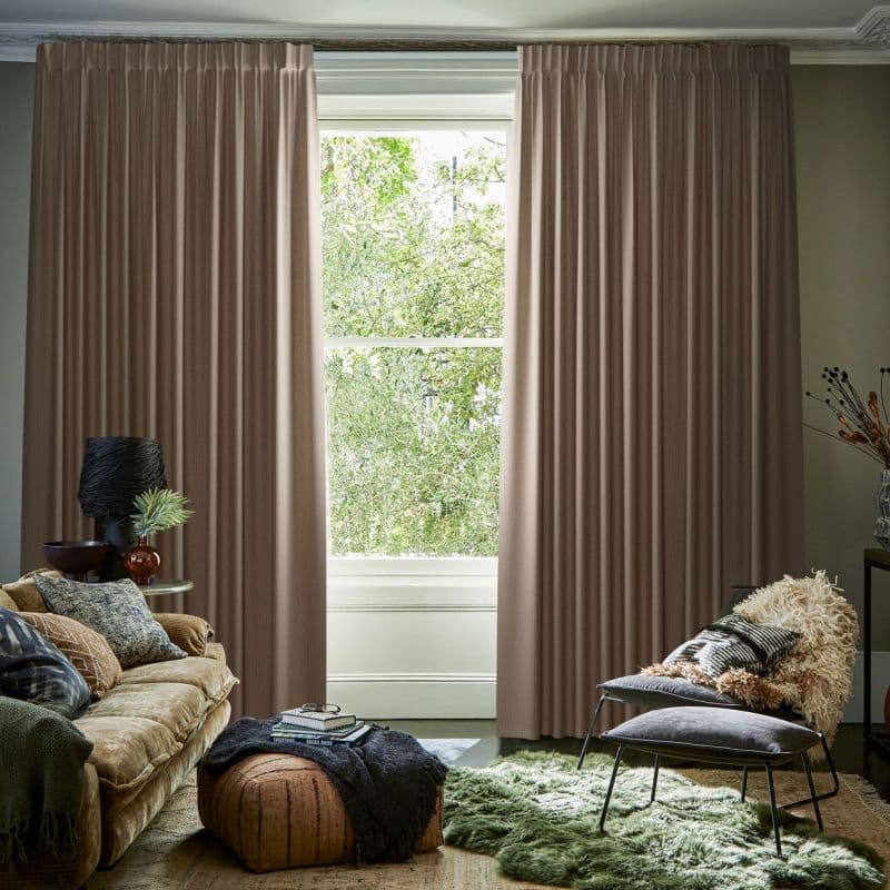 Full-length pleated beige curtains, hung on a ceiling track, mostly closed with a narrow central opening, framing and filtering daylight from a leafy garden visible through a living-room window.