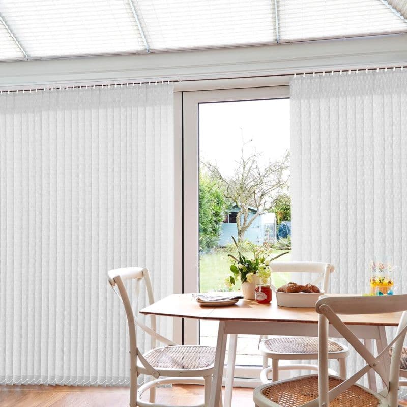 White vertical blinds mostly closed with a central gap exposing a glass patio door; filtering daylight into a dining area with table, chairs, and a backyard with tree.