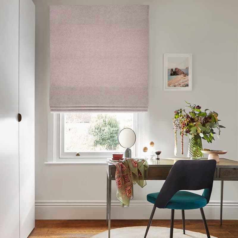 Soft pink fabric Roman blind, lowered to just above the sill and lightly filtering daylight, over a home desk with teal chair, vase of flowers and a small round mirror.