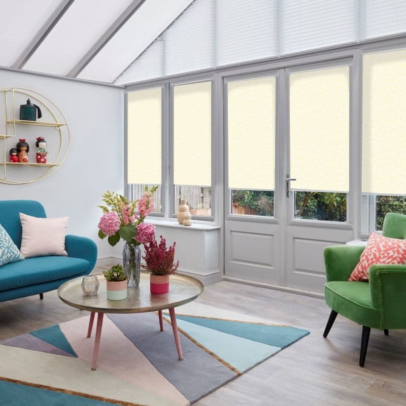 Pale roller blinds over tall windows and a French door, lowered across upper panes to soften daylight; bright conservatory living room with sofa, armchair, coffee table, flowers, and garden view.