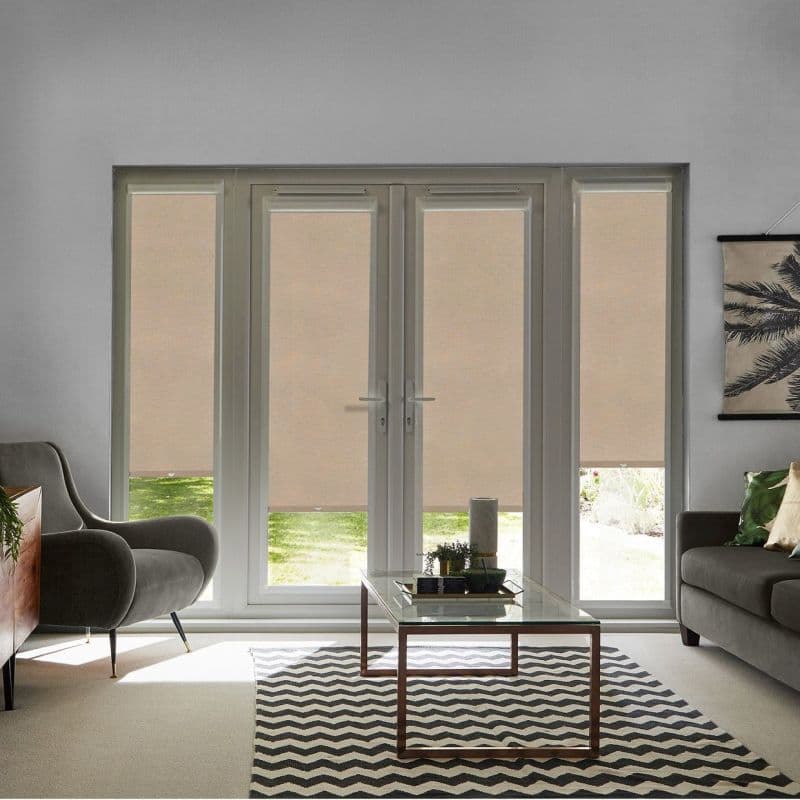 Beige roller blinds covering four tall French doors, lowered nearly to the sill and filtering daylight; living room with glass coffee table, chevron rug, armchairs and grassy garden view.