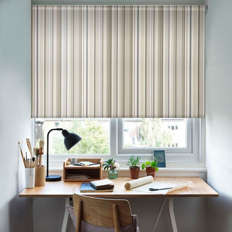 Striped beige-and-grey roller blind, lowered across a double window with vertical narrow stripes; filtering soft daylight into a small home workspace with wooden desk, lamp, art brushes and potted plants.