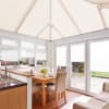 Cream pleated cellular blinds cover the pitched conservatory roof and windows, filtering bright daylight; in a light-filled breakfast/dining extension with wooden table, upholstered chairs, kitchen sink, and patio view.