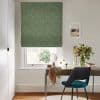 A green textured Roman blind, fully lowered with a slight bottom gap, filtering daylight into a home study; wooden desk, teal chair, round mirror, vase of flowers and framed seaside print.
