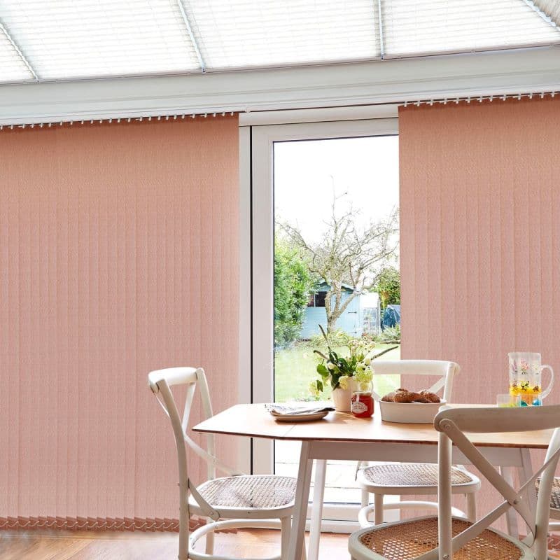 Peach vertical blinds (full‑length, narrow slats) cover a patio door, partly parted to let soft daylight through; a small dining table with white chairs, flowers, and pastries sits inside.