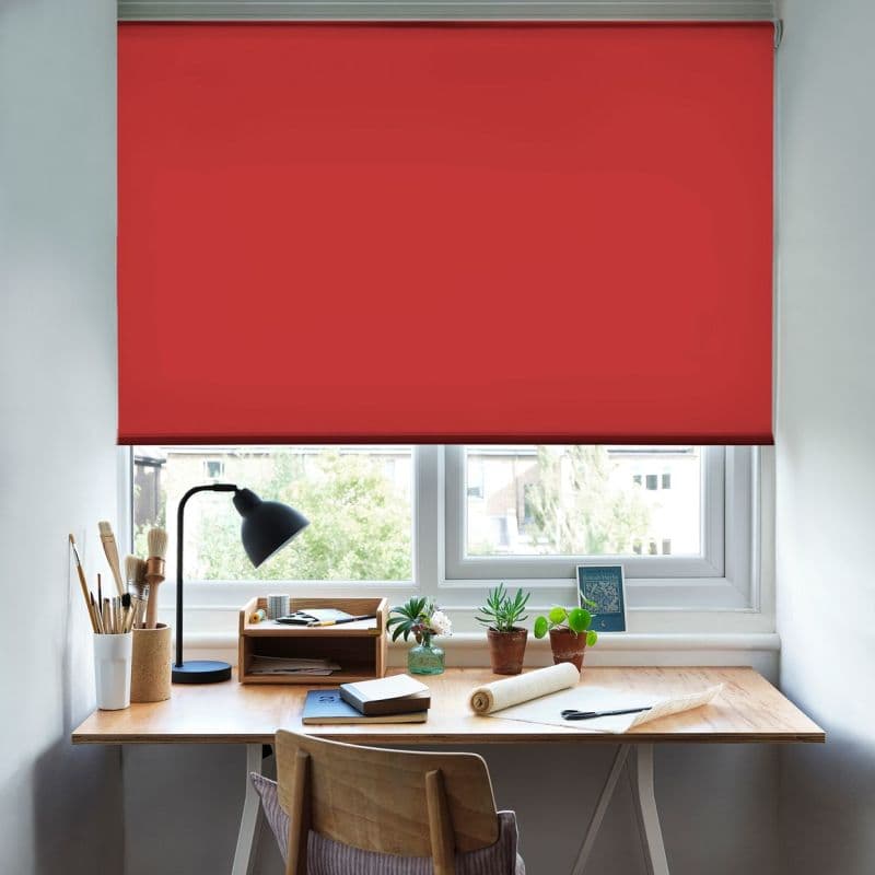 Red roller blind, lowered nearly to the sill, covering the window and filtering daylight over a tidy home desk with lamp, potted plants, notebooks, and a treed outside view.