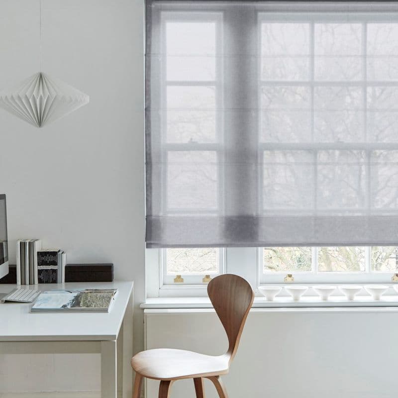 Sheer gray Roman shade (window dressing), lowered mid‑window and softly filtering daylight; simple white home office context with a wooden chair, desk, small pendant lamp and books.