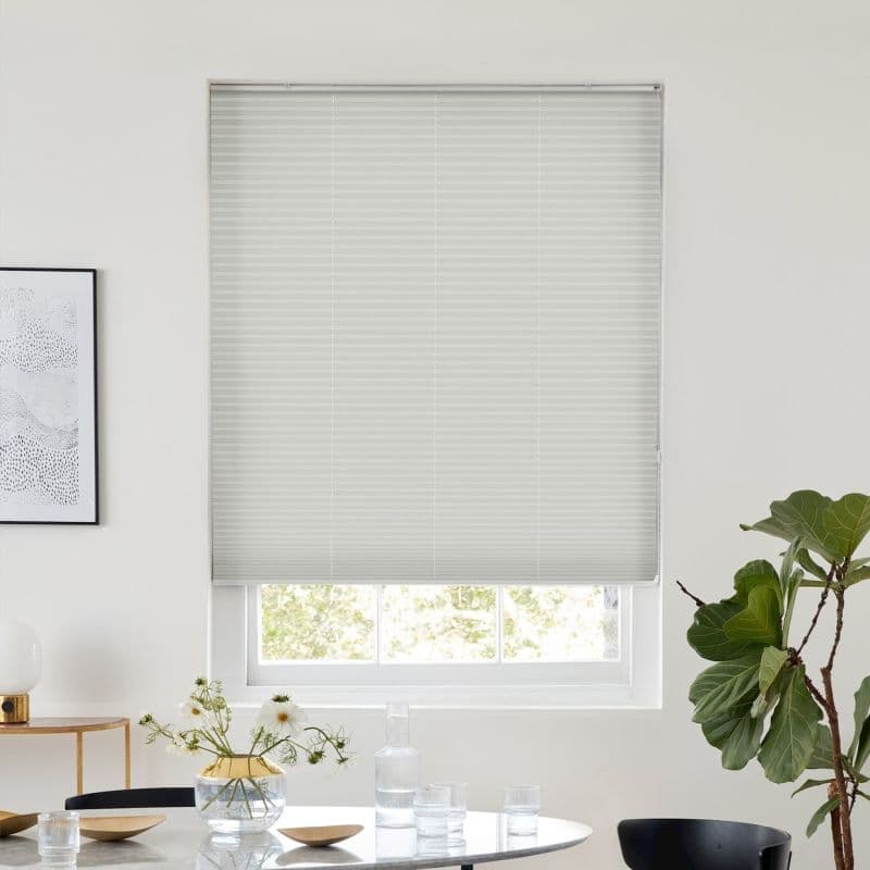 White cellular blind (honeycomb), lowered with horizontal pleats and slight lift at bottom, filtering daylight; over a window in a bright minimalist dining area with table, vase, and potted plant.