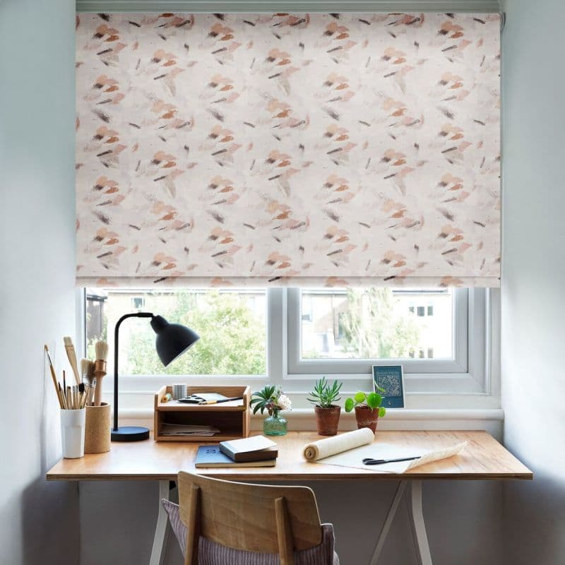 A pastel abstract-patterned roller blind lowered over a double window, gently filtering daylight; positioned above a wooden desk with lamp, notebooks, potted plants and a suburban leafy view.