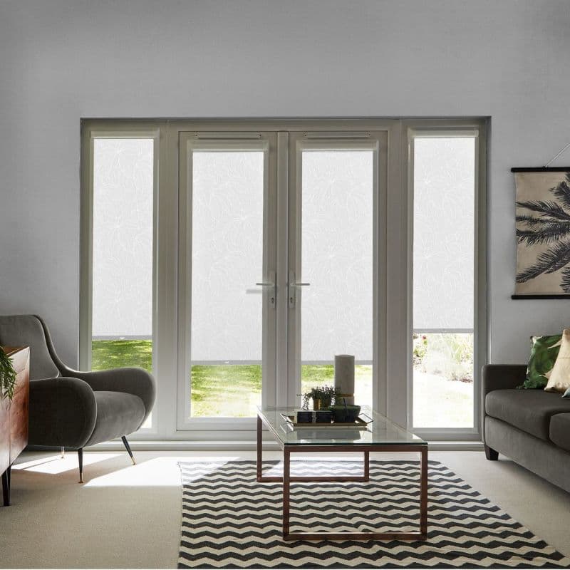 White patterned roller blinds covering French doors and sidelights, lowered to a narrow bottom gap, diffusing daylight and offering privacy; living room with grey sofa, table, rug, garden visible.