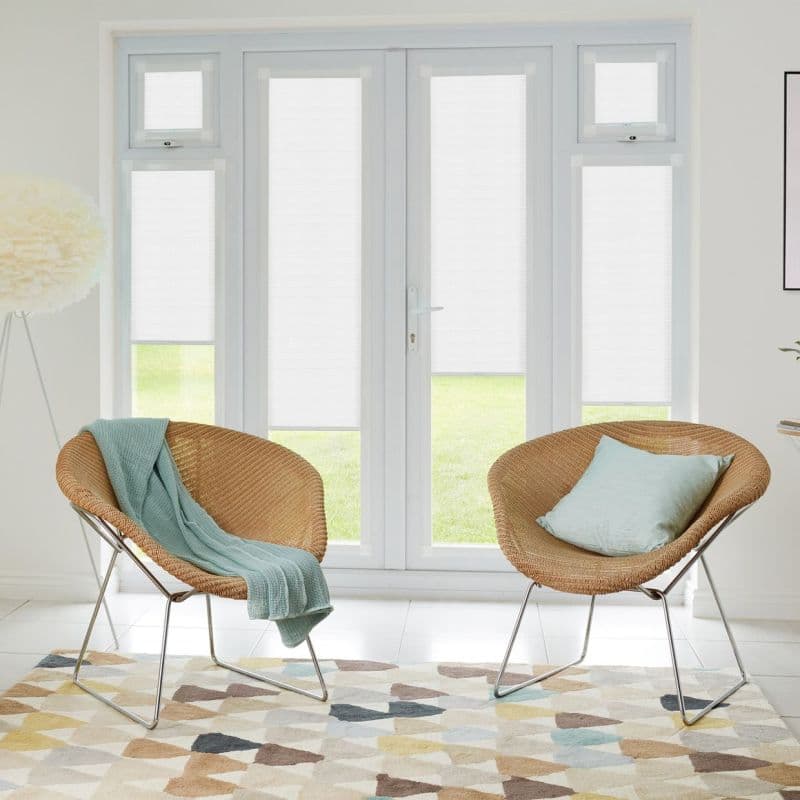 White pleated blinds fitted inside tall French doors and sidelights, closed and diffusing bright daylight; set in a neutral sitting area with two rattan chairs, pastel throw and patterned rug.