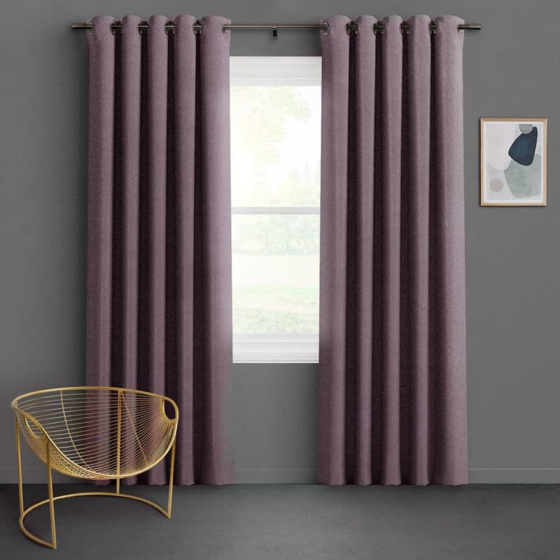 Floor-length grommet-top mauve curtains, hung on a metal rod, are parted to frame a sunlit window, filtering daylight in a grey-walled room with a gold wire chair and abstract art.