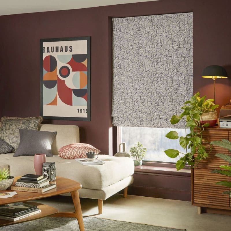 Roman blind lowered: white fabric with dense navy geometric line pattern, closed and softly diffusing light; in a plum-walled living room with a beige chaise, mid-century coffee table, plants, and poster.
BAUHAUS
1919
