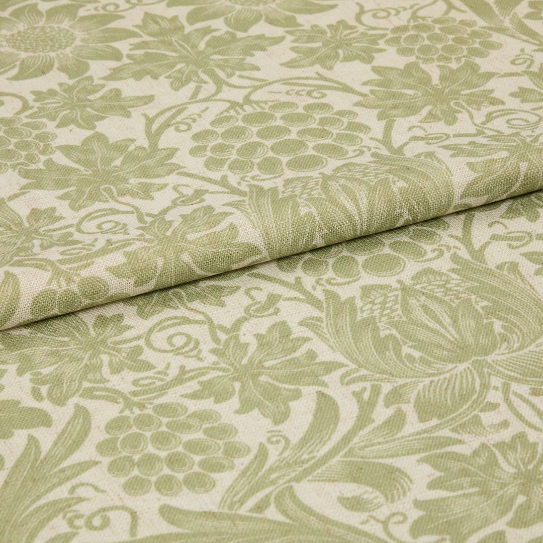Green-and-cream floral-patterned fabric, folded with an overlapping edge, displaying woven texture and grape-and-leaf motifs; close-up studio shot with soft, even lighting suggesting an upholstery or curtain sample.