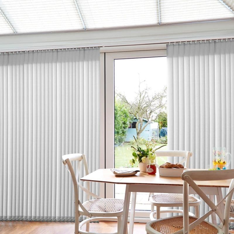 White vertical blinds cover a sliding glass door, slats mostly closed with a central gap, filtering daylight into a dining nook with wooden table, chairs, vase, and a backyard view.