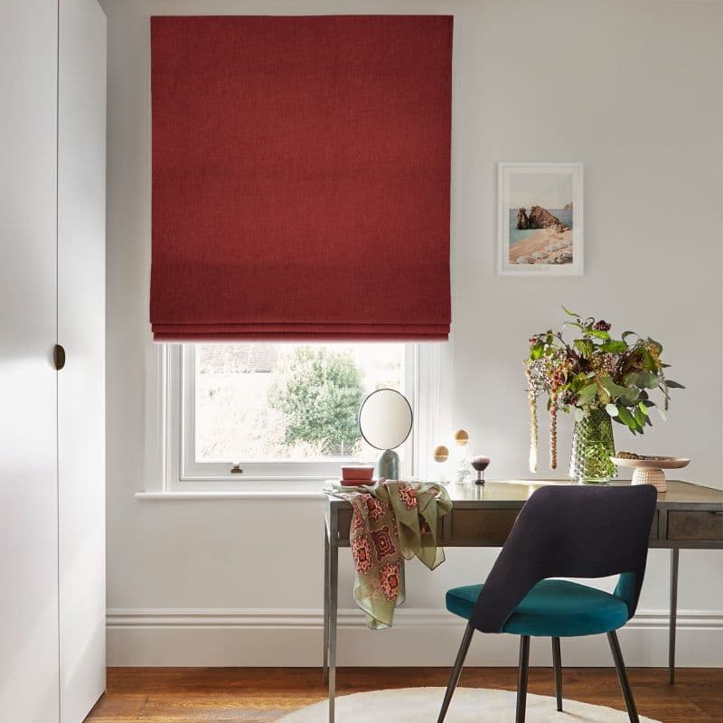 Red fabric Roman blind, lowered with soft folds, filtering daylight through a narrow lower gap; in a bright office with desk, teal chair, vase of flowers and framed coastal print.