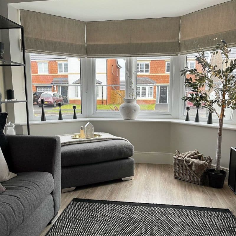 Beige fabric Roman blinds lowered across a three‑panel bay window, softly filtering daylight; decorative vase, candlesticks, potted tree and grey sofa/ottoman in a bright suburban living room.