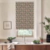 Patterned roller blind covers a window above a stainless-steel kitchen sink; geometric multicolored design contrasts with light walls, wood cabinets, faucet, potted plants, and framed sketch on countertop.