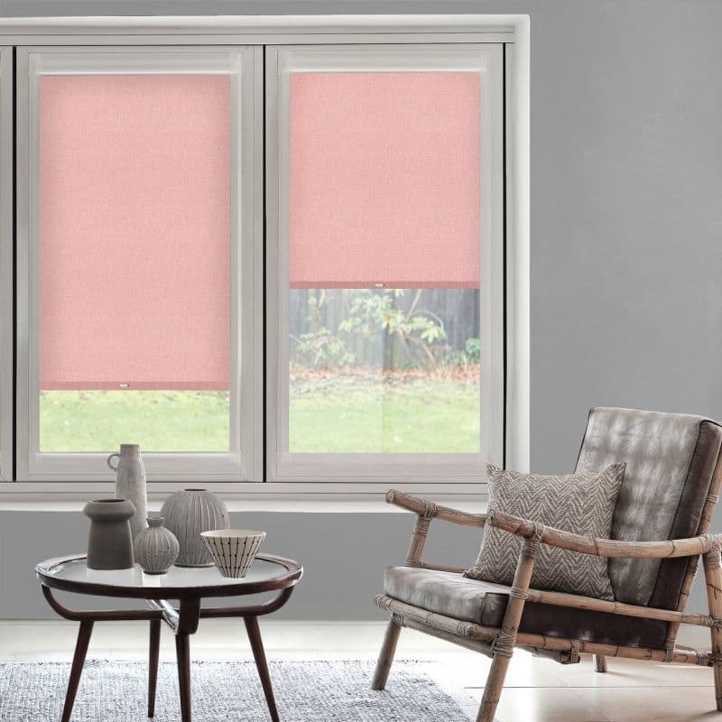 Pink fabric roller blinds on twin windows — left lowered, right raised halfway — filtering light in living room with rattan armchair, table with ceramics and a grassy yard outside.