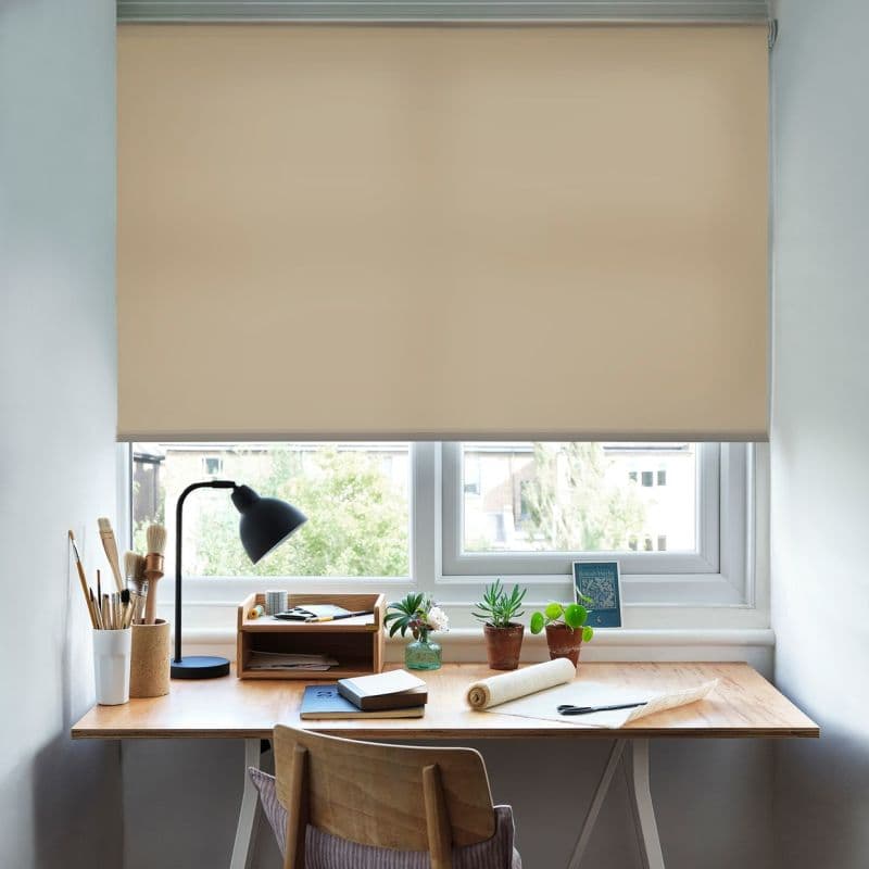 Beige roller blind, lowered to cover most of a double window, filtering light; wooden desk beneath holds a black desk lamp, potted succulents, brushes and notebooks in a bright workspace.