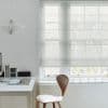 Sheer white Roman shade with horizontal folds, lowered over a multi-pane window, filtering soft daylight; minimalist home office with white desk, wooden chair, pendant lamp and bare tree branches outside.