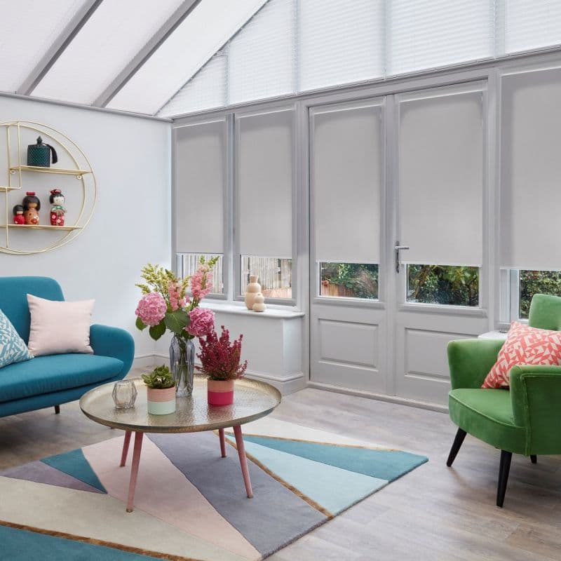 Grey roller blinds lowered across tall glazed doors and upper panes, mostly closed and filtering daylight with narrow bottom gaps; conservatory living area with colorful sofa, armchair, coffee table, flowers.