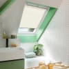 Cream pleated skylight blind, nearly lowered over an angled roof window, softly filtering daylight. Bathroom setting with green window surround, white herringbone tiles, bathtub, sink, plant and bath accessories.