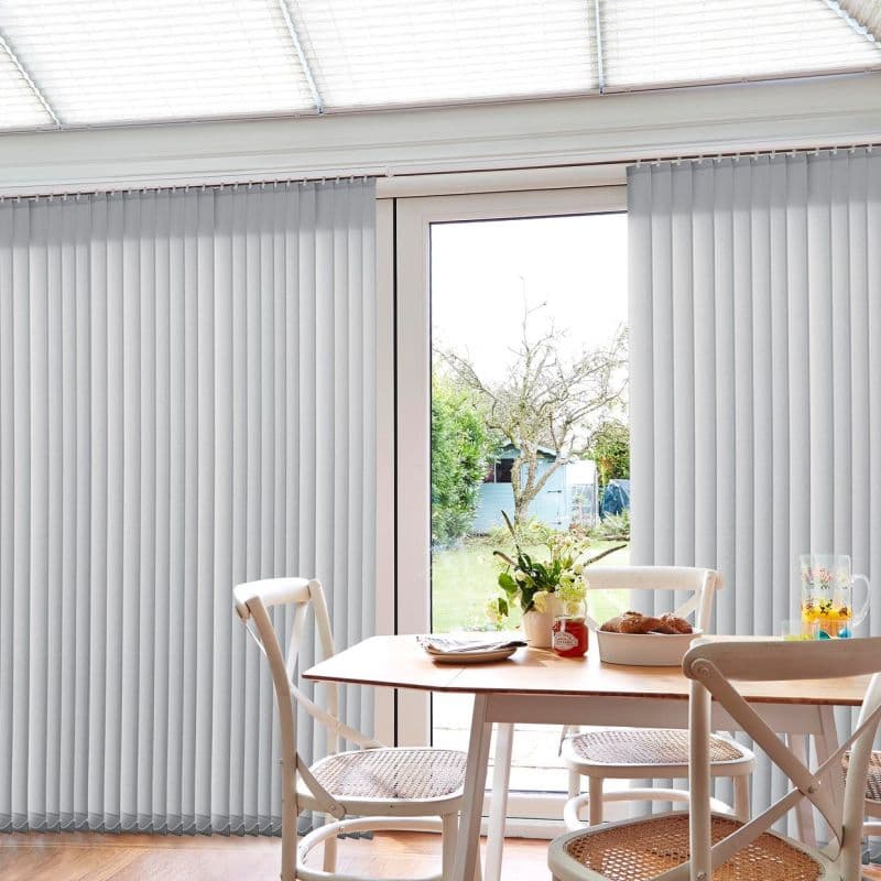 Vertical pale-grey fabric blinds cover a patio door, closed with narrow slats filtering light; dining table and woven chairs in foreground, soft daylight and garden visible through a central gap.