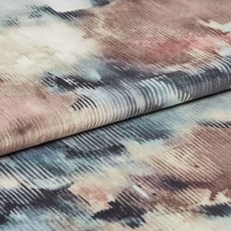 Folded ribbed velvet fabric with watercolor-like washes of blue, pink, beige and black — layered to reveal horizontal ribs and soft pile — close-up on a softly lit neutral surface.