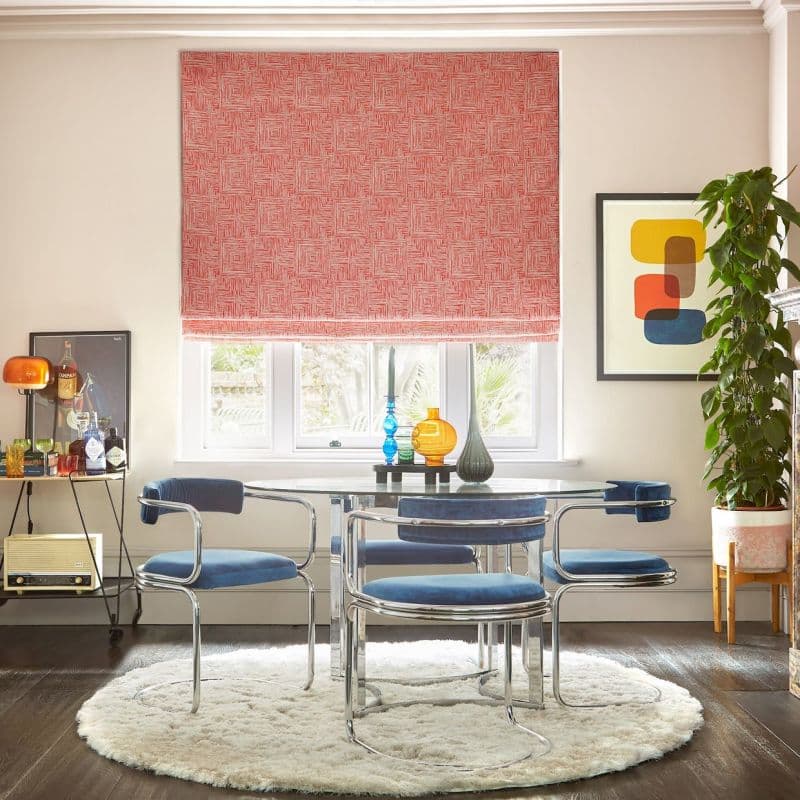 Red patterned Roman blind lowered over a three‑pane window, filtering daylight; glass dining table with chrome‑framed blue chairs, round shag rug, potted plant and colorful vases; garden view. Campari