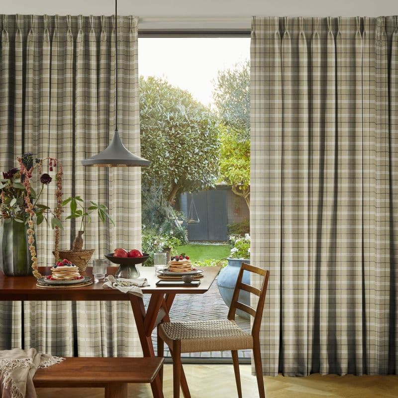 Beige plaid pleated curtains, mostly drawn with a central gap revealing the outside; filtering soft morning light over a wooden dining table set for breakfast, pendant lamp, and garden view.