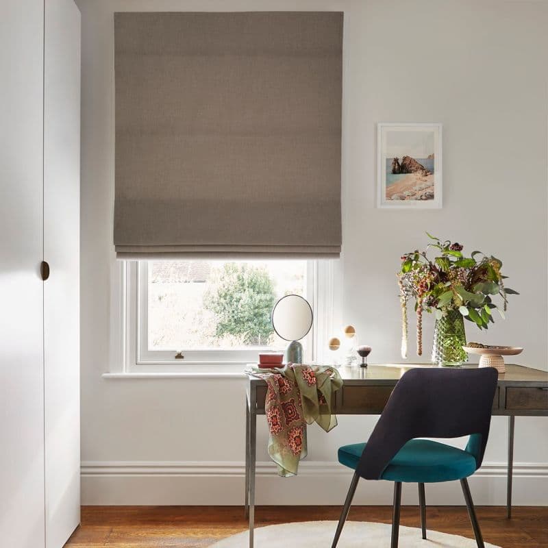 Taupe Roman blind, mostly lowered with soft folds, filtering daylight through a narrow gap; over a desk in a bright home office with vase of flowers, mirror and teal chair.