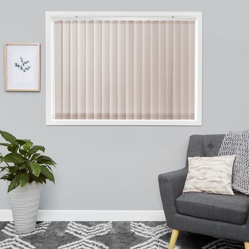 Beige vertical blinds covering a centered rectangular window, closed and gently diffusing light; set on a grey living-room wall with a charcoal armchair, knitted throw, potted plant, and patterned rug.
