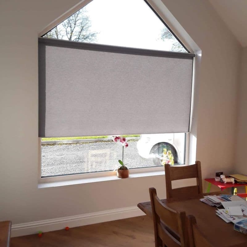 A grey roller blind, lowered to cover most of a tall triangular‑top window, filtering light; dining area with wooden table and chair, small potted orchid on the sill, car visible outside.