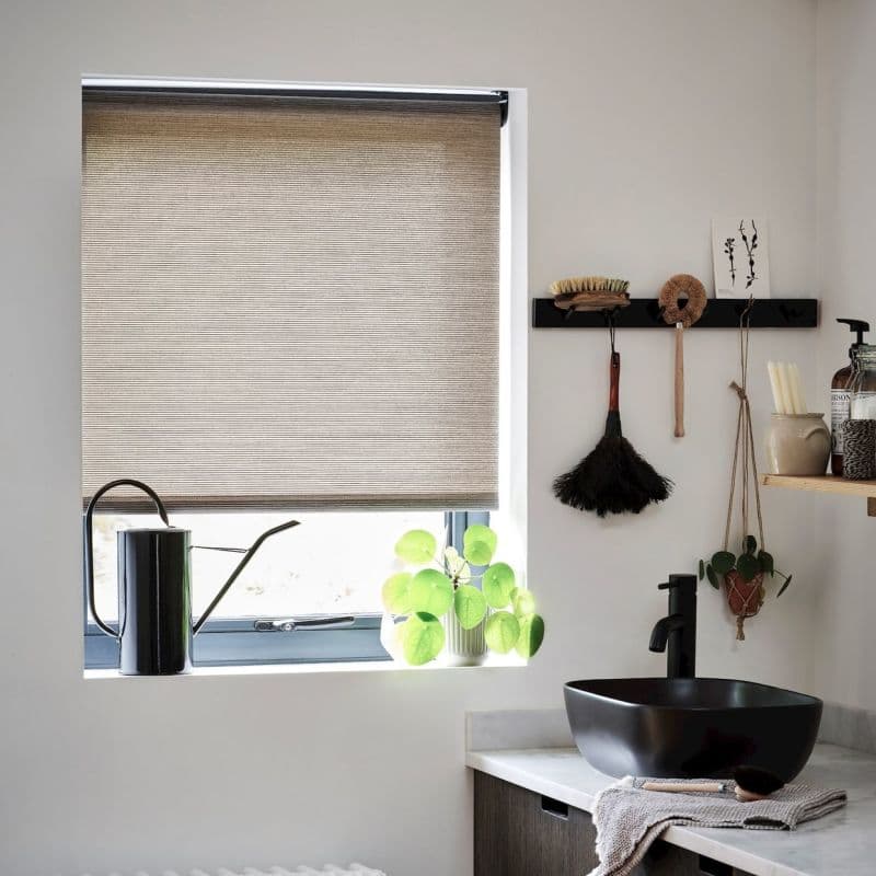 Beige fabric roller blind lowered over a recessed window, filtering daylight; foreground shows a black watering can, potted round‑leaf plant, black vessel sink and hanging brushes on a white wall.