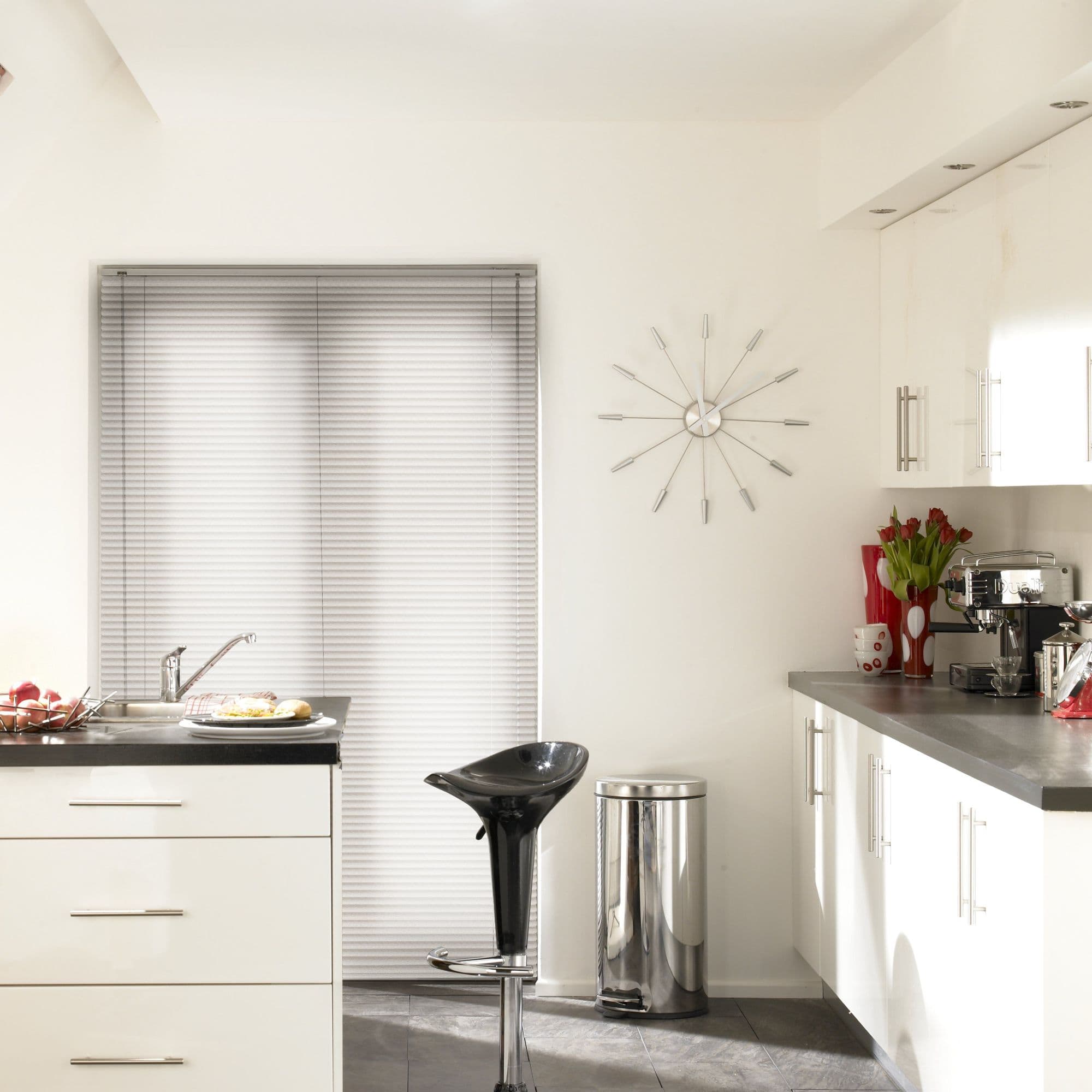 Horizontal aluminum Venetian blinds covering a doorway/window, fully closed and softly diffusing daylight; modern white kitchen with black bar stool, stainless bin, wall clock and countertop appliances.