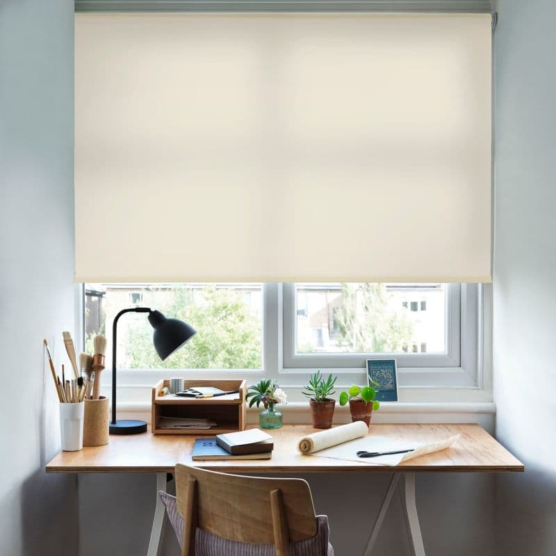 Cream roller blind, partially lowered and translucent, filtering daylight; mounted over a double window above a wooden desk with lamp, brushes, notebooks and potted succulents in a bright workspace.