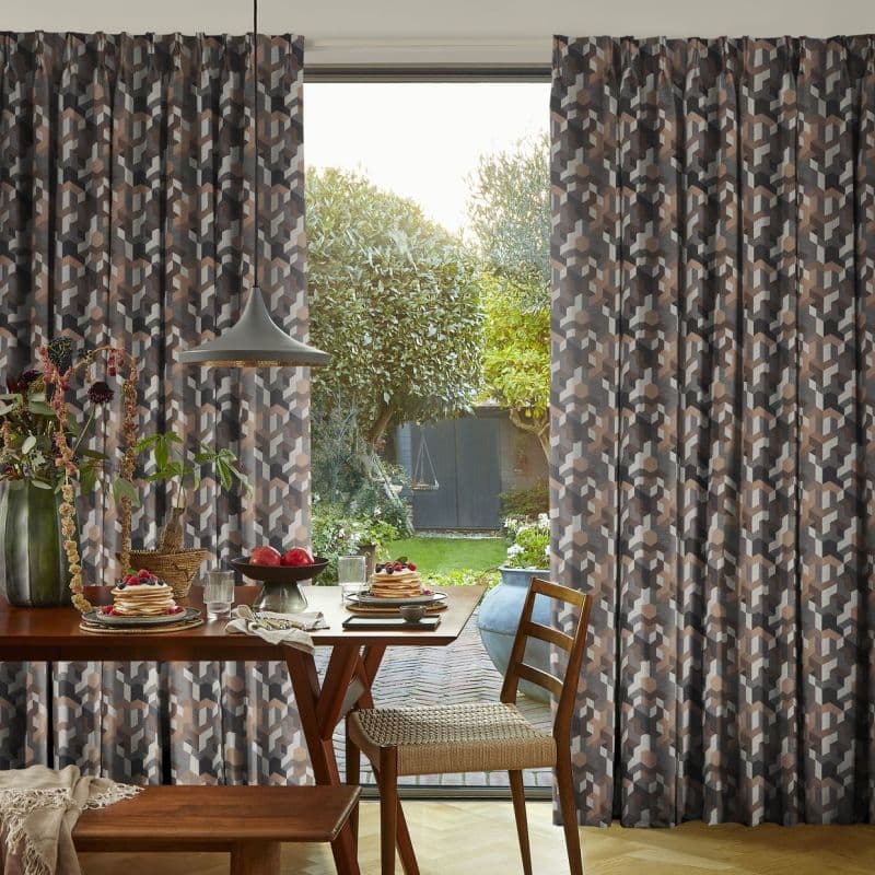 Geometric-patterned pleated curtains, full-length and drawn partly aside revealing a central gap; framing a sunlit backyard with manicured lawn, trees, patio and a breakfast table set inside by the window.