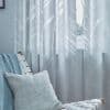 Sheer light-blue curtains with white abstract tree/leaf pattern hang closed over a window, filtering daylight; nearby a woven white chair holds a blue knit throw and pale cushion in a calm, lit room.