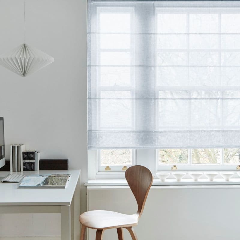 Sheer white Roman blind lowered over a multi-pane window, softly filtering daylight; minimalist home office with white desk, wooden chair, folded-paper pendant, books and small white bowls on the sill.