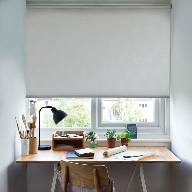 White fabric roller blind pulled down to cover most of a double-pane window, softly filtering daylight onto a wooden desk with lamp, brushes, potted succulents and stationery, suburban view below.