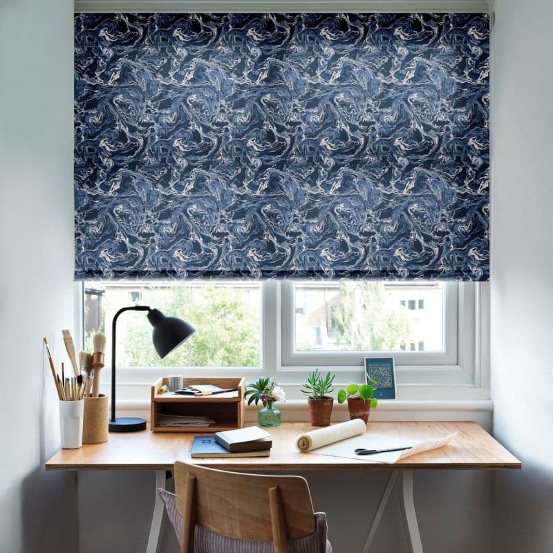 Roller blind: navy-blue marble swirl pattern, lowered halfway across a two-pane window — filtering daylight and blocking view; home study desk beneath, lamp, plants, art supplies on wooden surface.