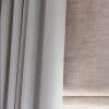 Grey pleated blackout curtain at left and beige textured Roman blind at right; both lowered, the curtain drawn closed and the blind filtering diffuse daylight in a close-up interior view.