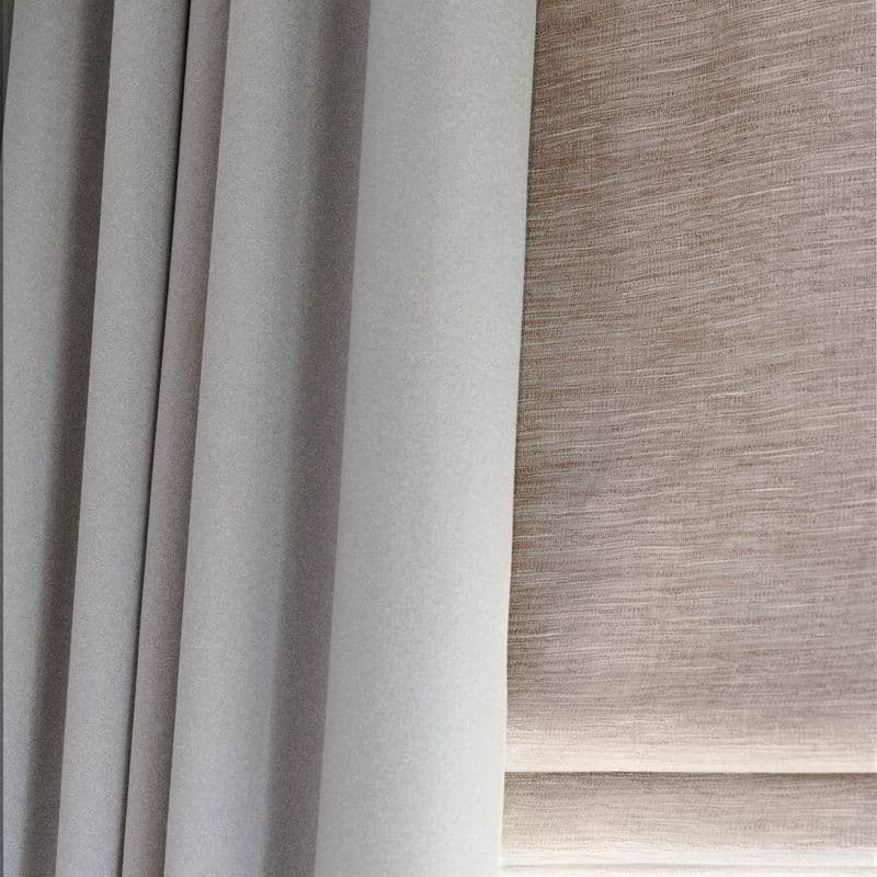 Grey pleated blackout curtain at left and beige textured Roman blind at right; both lowered, the curtain drawn closed and the blind filtering diffuse daylight in a close-up interior view.